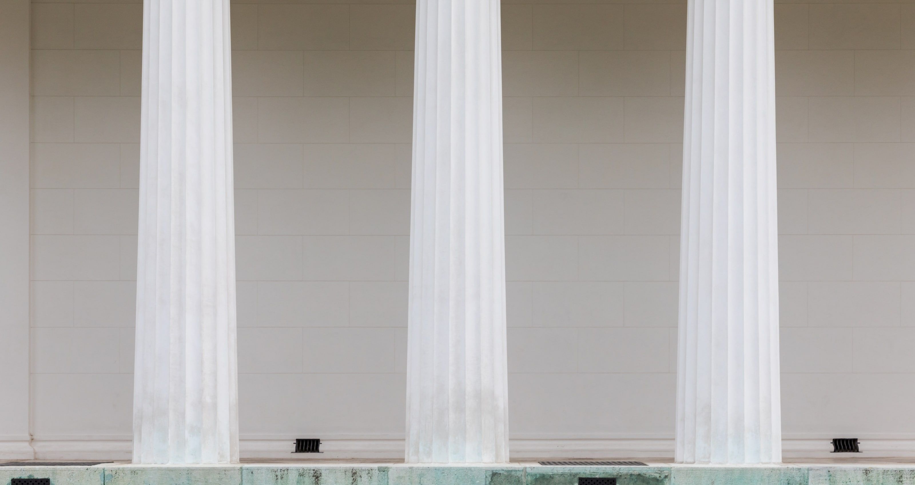 Three white fluted Doric columns standing in front of a pale stone wall.